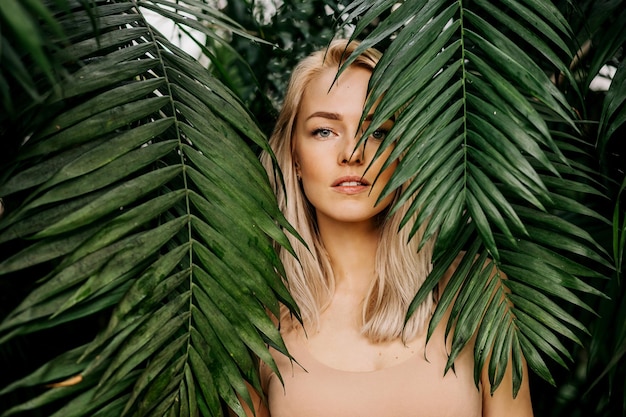 Young woman's face surrounded by tropical leaves. Closeup portrait of magnificent lady with natural nude makeup, perfect skin, blonde hair. Natural cosmetic, wellness, purity, skincare, beauty concept