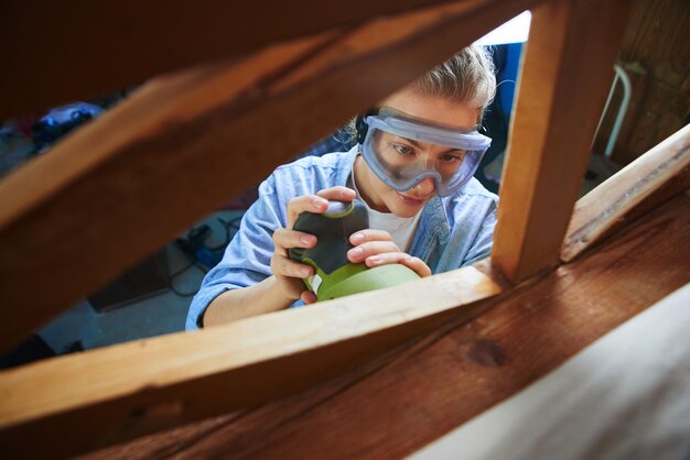 Young woman in safety glasses polishes a wooden bed with random orbit sander, diy concept