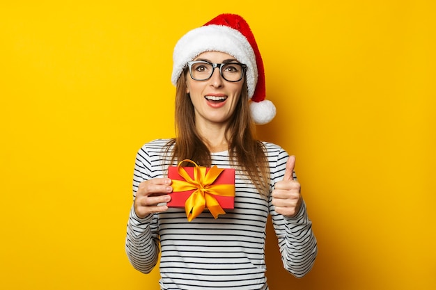 サンタクロースの帽子をかぶった若い女性がクリスマスプレゼントを持っています