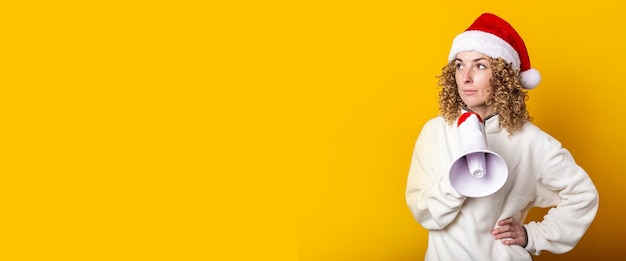 Young woman in santa claus hat with a megaphone on a yellow background Banner