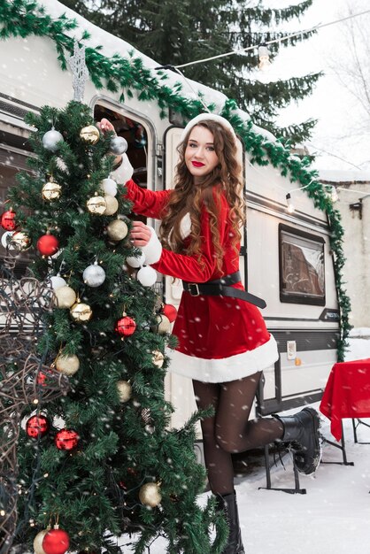Young woman in santa costume decorates the christmas tree at winter campsite getting ready for the