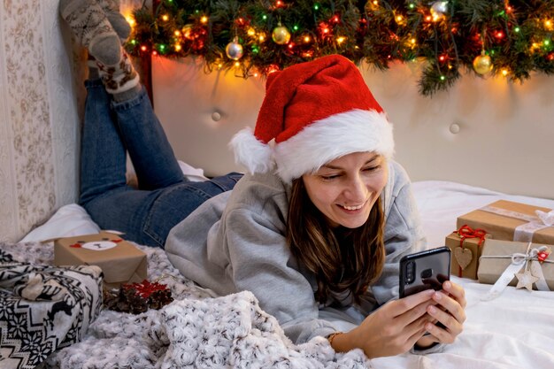 サンタの帽子をかぶった若い女性は、電話でオンラインで家族や友人を祝福します