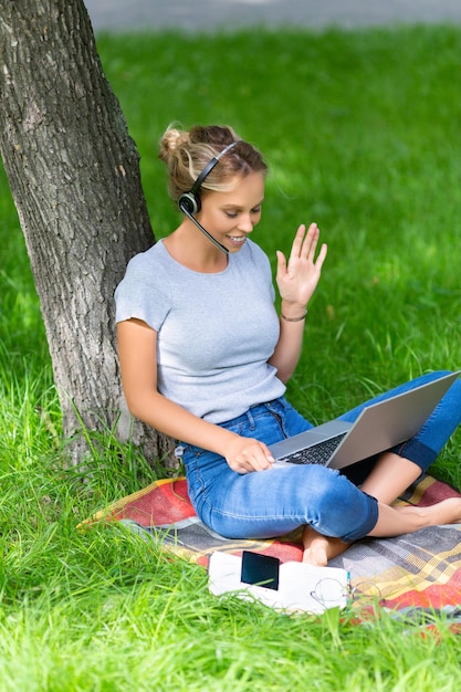 Молодая женщина здоровается по видеосвязи с коллегами на онлайн-брифинге ноутбука, сидя в парке