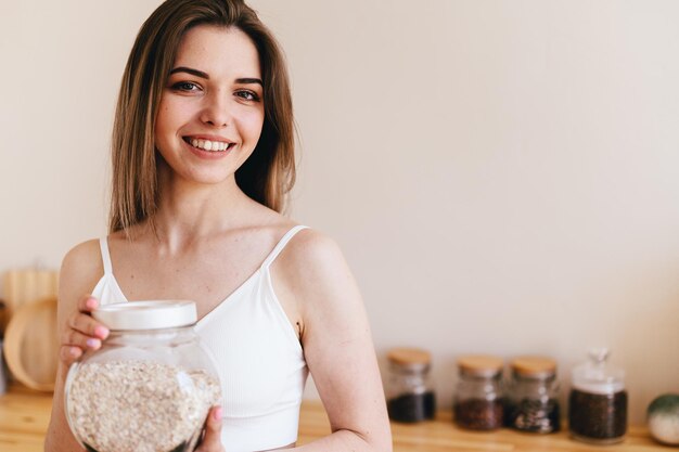 Young Woman Scratches her Head and Looks out Window Girl Holds in her Hands Glass jar with Oatmeal