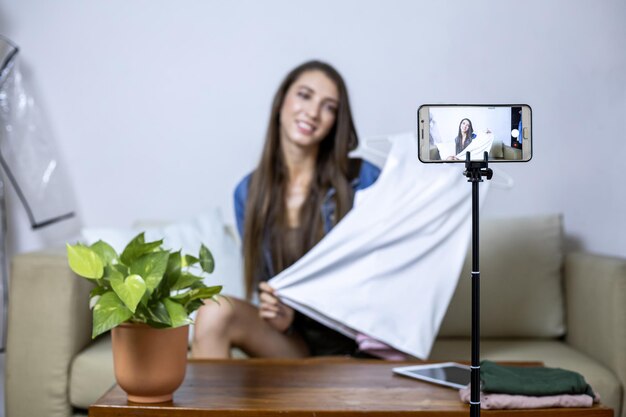 Young woman selling clothes online via her mobile phone at home. Sell stuff online. Live selling.