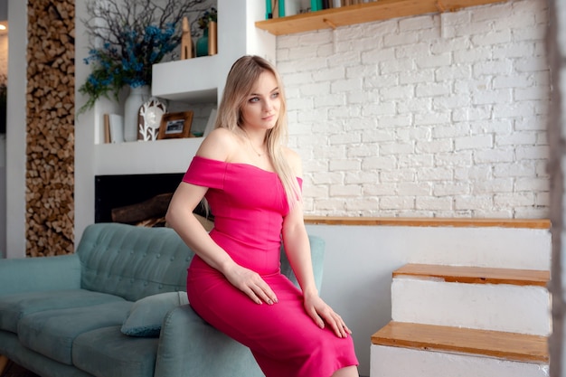 Young woman in sexy dress is sitting on a couch. Sensual beautiful blonde woman posing in pink dress. Girl with long hair. Indoor shot.