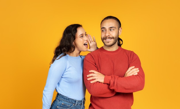 Young woman sharing secret with her husband yellow background