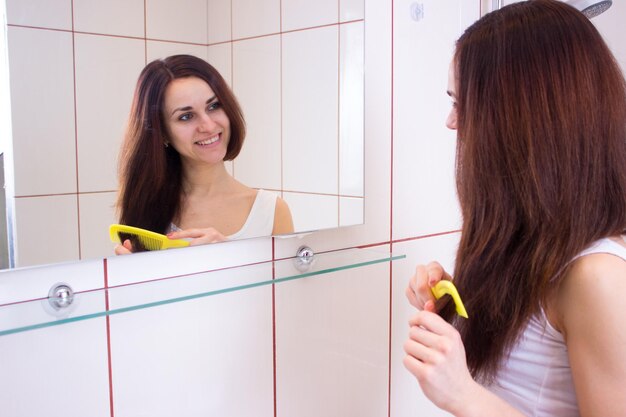 셔츠를 입은 젊은 여성이 욕실 거울 앞에서 긴 갈색 머리를 빗으로 빗고 있다