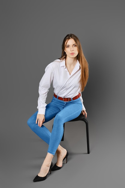 Foto una giovane donna in camicia e jeans si siede su una sedia in una postura tesa su uno sfondo grigio dello studio