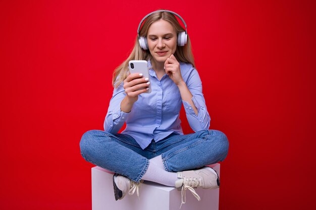 A young woman in a shirt listens to music from her phone on a red background