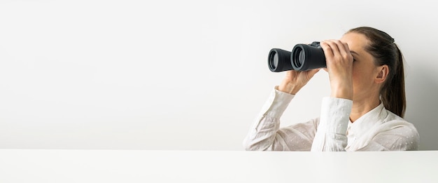 Young woman in shirt looking through binoculars looking out from behind cardboard Banner