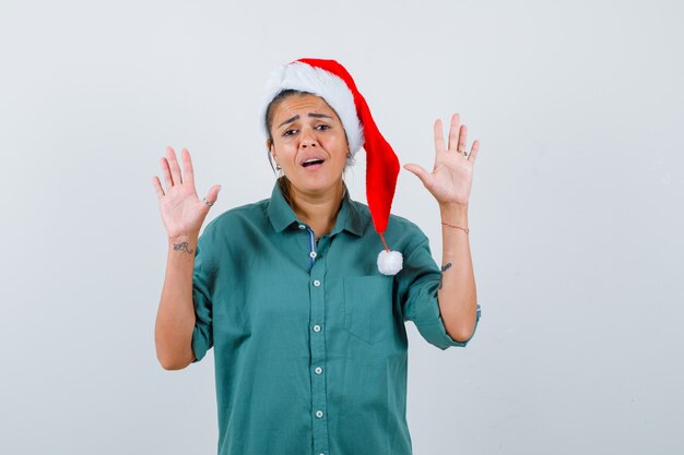 Young woman in shirt, Santa hat showing surrender gesture and looking helpless , front view.