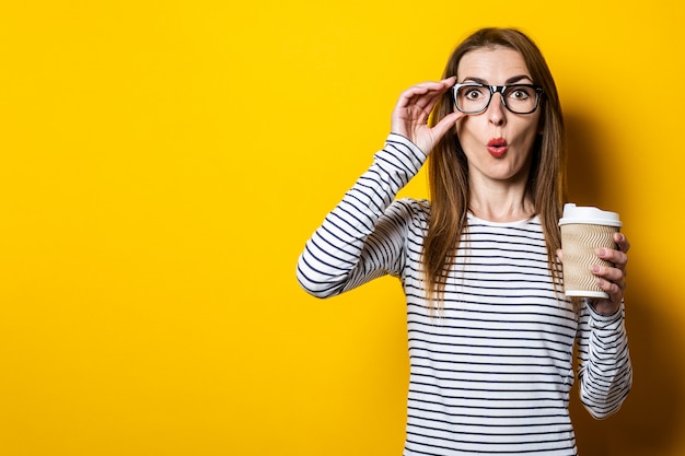 Young woman shocked in glasses with a paper cup of coffee on a yellow background