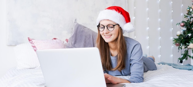 크리스마스를 위해 온라인으로 쇼핑하는 젊은 여성은 파란색 아늑한 옷을 입고 침대에 앉아 있다