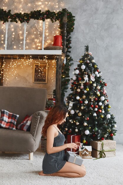 A young woman in a short dress unpacking a gift box and sits on the floor near the Christmas tree