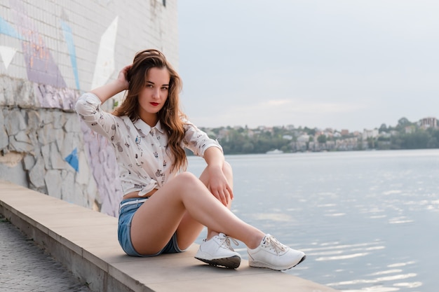 Young woman in shorts and sneakers sitting on the background of the lake. Beautiful girl posing on the background of the lake on a sunny day. Girl in casual clothes