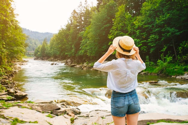 young Woman in shorts and straw hat standing on the rocks near the river