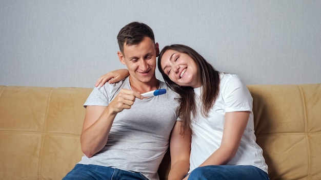 Young woman showed a pregnancy test to a man sitting on the couch. The husband is very happy to become a father.