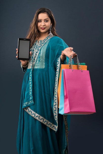 Young woman showing blank screen of mobile phone or tablet smartphone and shopping bags in her hands on a grey background