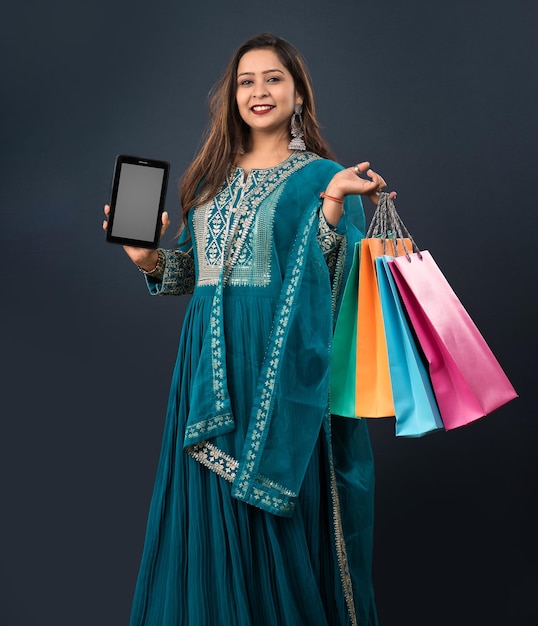 Young woman showing blank screen of mobile phone or tablet smartphone and shopping bags in her hands on a grey background