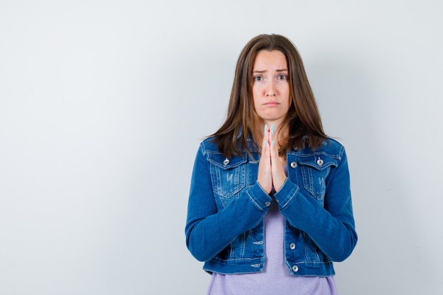 Young woman showing clasped hands in pleading gesture in denim jacket and looking dismal , front view.