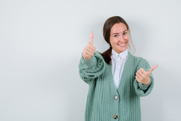 Young woman showing double thumbs up in blouse, cardigan and looking merry , front view.