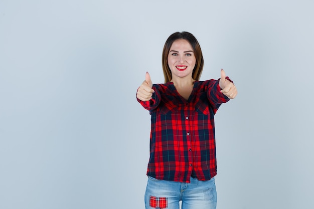 Молодая женщина показывает двойные пальцы вверх в клетчатой рубашке и выглядит радостной, вид спереди.