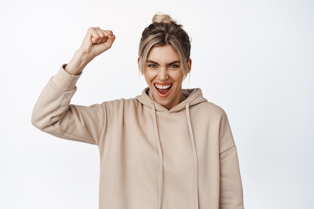 Young woman showing endorsement shaking fist and shouting rooting for team watching sports game standing against white background