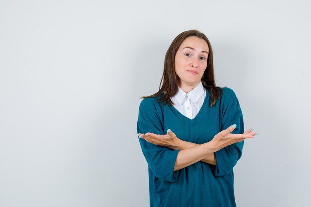 Giovane donna che mostra gesto impotente in maglione sopra una camicia bianca e sembra confusa, vista frontale.