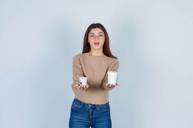 Young woman showing paper cups in sweater, jeans and looking curious, front view.