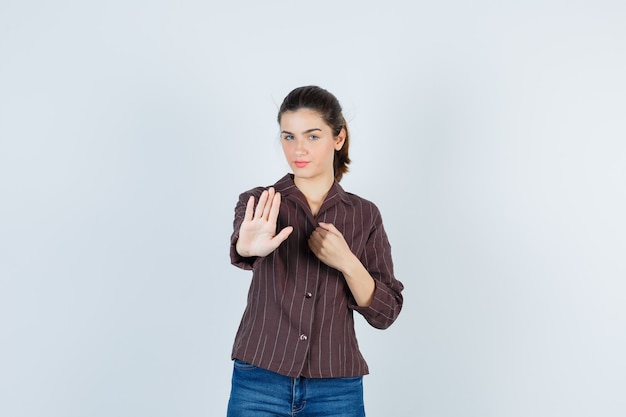 Young woman showing stop sign, holding fist over chest in striped shirt, jeans and looking serious. front view.