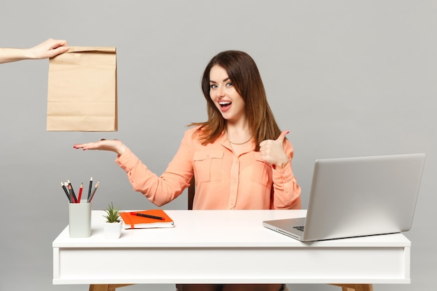 Young woman showing thumb up pointing hand on brown clear empty\
blank craft paper bag for takeaway work at desk with laptop\
isolated on gray background. achievement business career lifestyle\
concept.