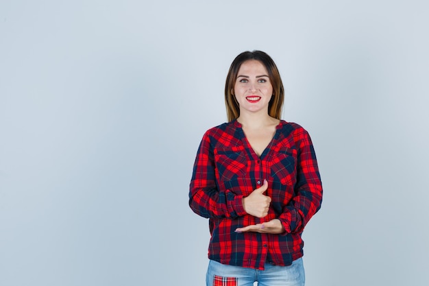 Young woman showing thumb up, spreading palm out in checked shirt and looking jolly , front view.