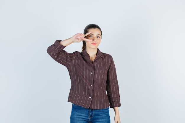 Young woman showing v sign on eye in striped shirt, jeans and looking happy. front view.