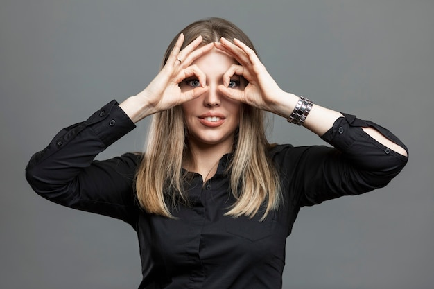 Young woman shows ok sign by closing her eyes. Masonic signs and symbols, the theory of the world conspiracy. Beautiful blonde in a black shirt. Gray background.