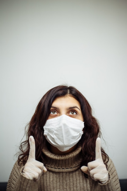 Young woman shows up sign with her hands and has to stay home during quarantine due to coronavirus pandemia. Beautiful girl sitting in flat wearing medical mask and gloves. Covid-19 epidemia concept.