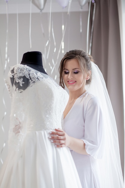 Young woman in silk bathrobe touching her wedding dress and smiling