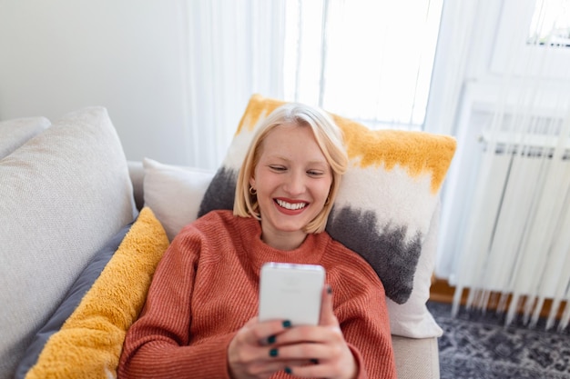 Молодая женщина отдыхает на диване в гостиной, просматривая беспроводной интернет на смартфоне. Тысячелетняя девушка отдыхает на диване дома. Текст сообщения на современном мобильном телефоне.