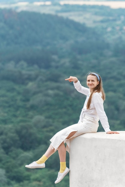 Young woman sits on balcony with her legs hanging down and points into the distance on mountains background Vertical frame