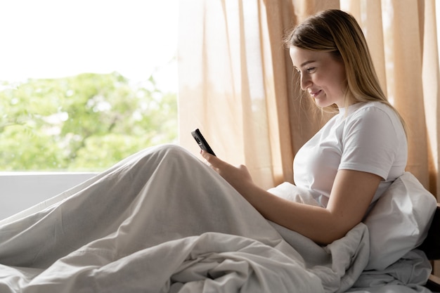 A young woman sits on a bed early in the morning. Reads great news on the phone.