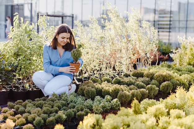 A young woman sits by a flower bed on the street near a plant shop and chooses a pot with a small tree A woman chooses plants for landscaping the yard Home gardening concept