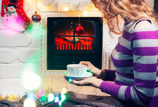 A young woman sits in front of the Christmas fireplace and holds a cup of tea in her hands