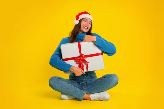 Young woman sits holding christmas gift box against yellow background