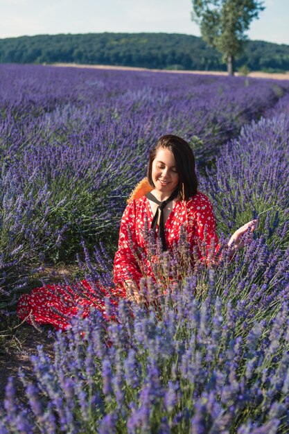 A young woman sits in a lavender field with a straw hat hanging around her neck