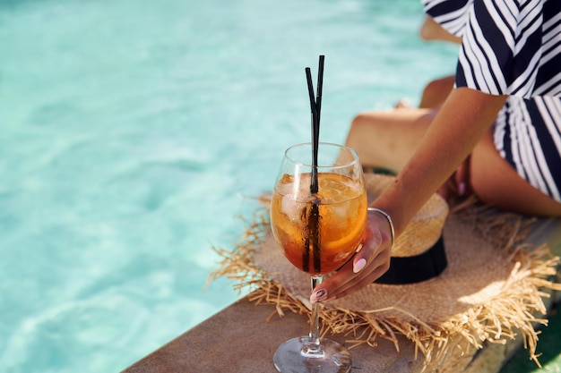 Young woman sits near swimming pool at daytime with cocktail