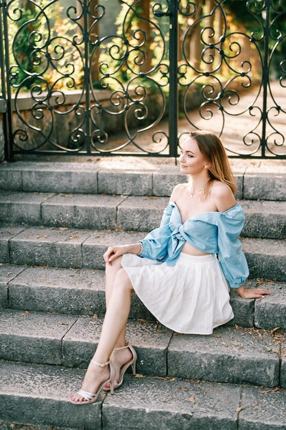 Young woman sits on stone steps in a garden near a forged fence and looks into the distance