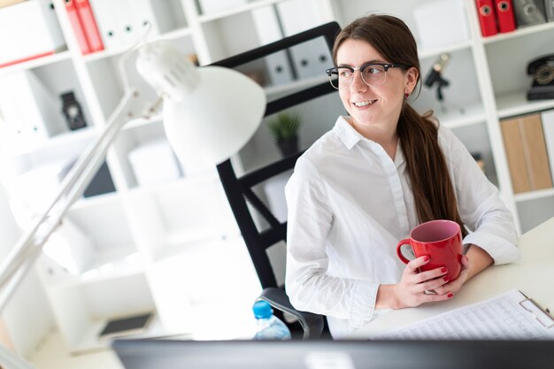 A young woman sits at a table in the office and holds a red cup in her hands.