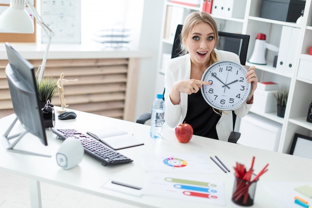 Una giovane donna si siede a un tavolo in ufficio e punta un dito sul quadrante dell'orologio.