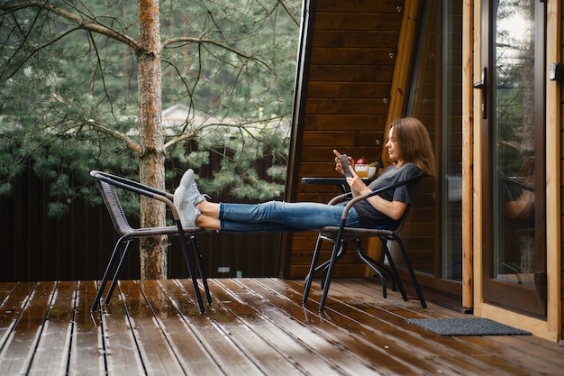Молодая женщина сидит на террасе каркасного дома с мобильным телефоном