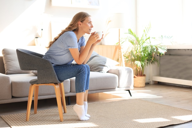 肘掛け椅子に座って、コーヒーを飲む若い女性。